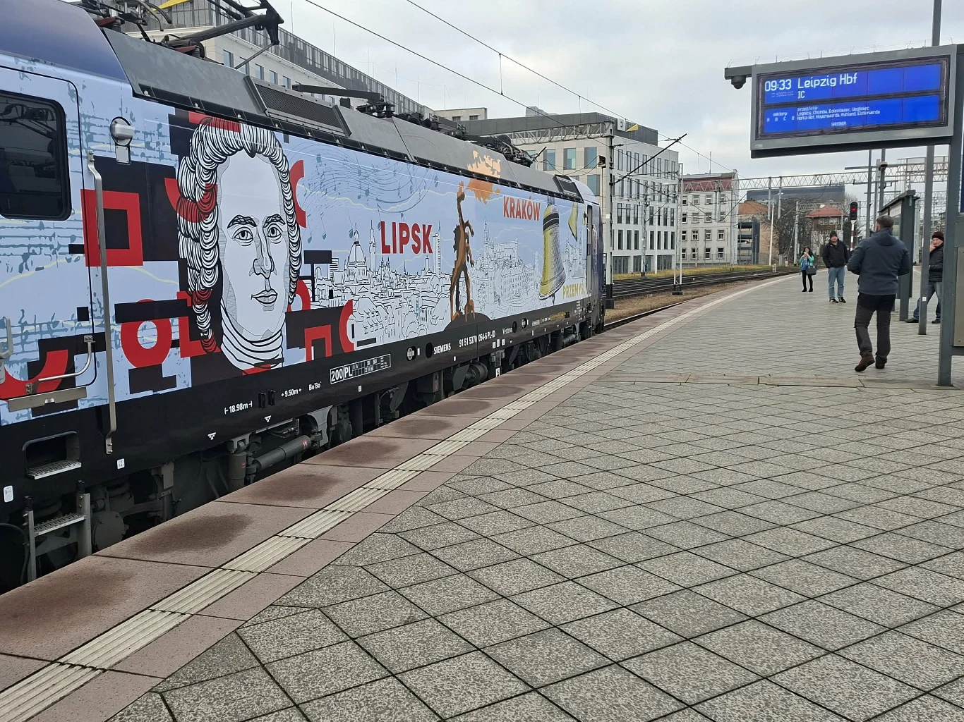 Okolicznościowa okleina lokomotywy na Wrocławiu Głównym Pociąg ozdobiony wielkoformatową grafiką, w tym portretem postaci historycznej oraz napisami 'Lipsk' i 'Kraków', stoi na peronie nowoczesnej stacji kolejowej. W tle widoczne budynki miejskie, kilku przechodniów oraz elektroniczny rozkład jazdy.