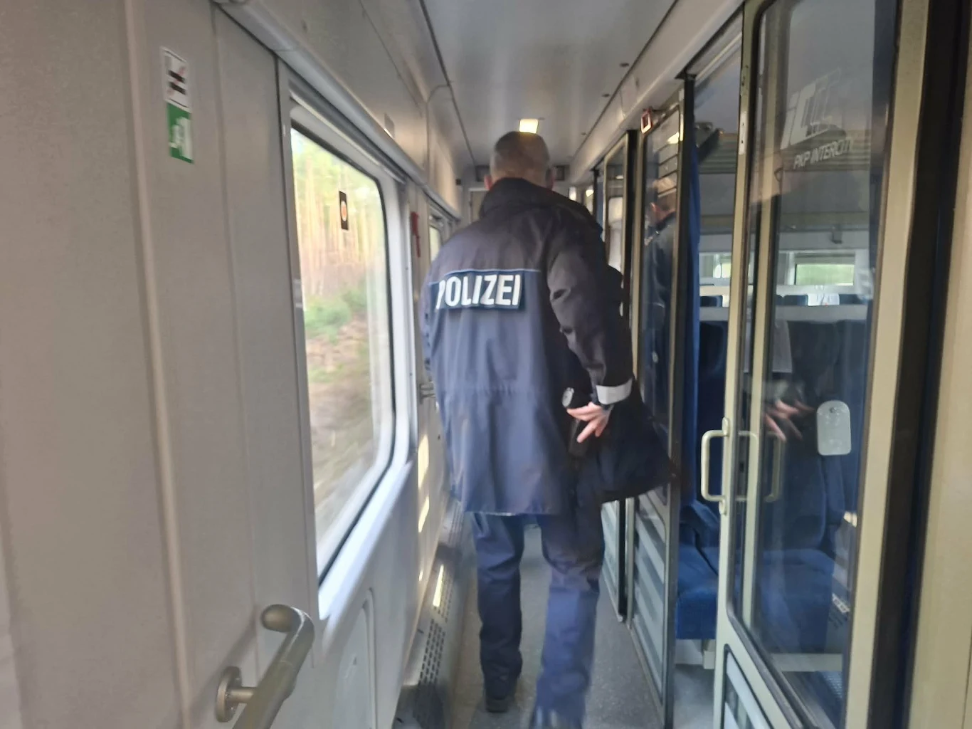 Niemiecki policjant na kontroli w pociągu do Lipska Funkcjonariusz policji w mundurze przechodzi przez wąski korytarz pociągu, po jego lewej stronie znajduje się okno, po prawej przedziały pasażerskie z przeszklonymi drzwiami.