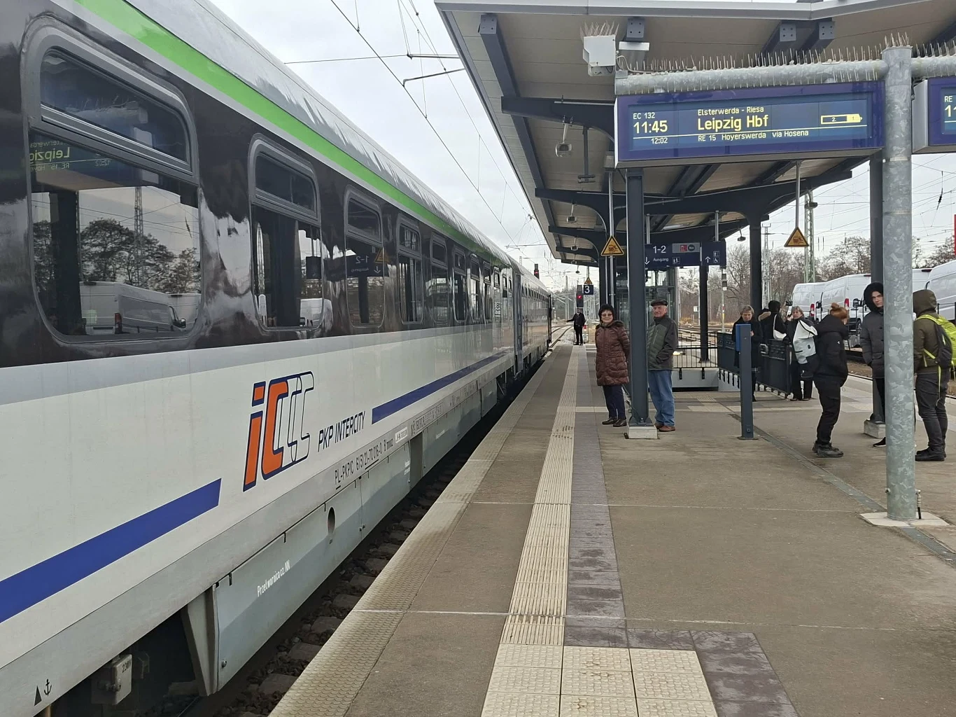 Pociąg na stacji kolejowej Ruhla Pociąg pasażerski IC stoi na peronie stacji kolejowej, kilka osób oczekuje przy krawędzi peronu, osłonięci ciepłymi ubraniami, widoczne są elementy infrastruktury kolejowej oraz tablica informacyjna z godziną odjazdu pociągu.