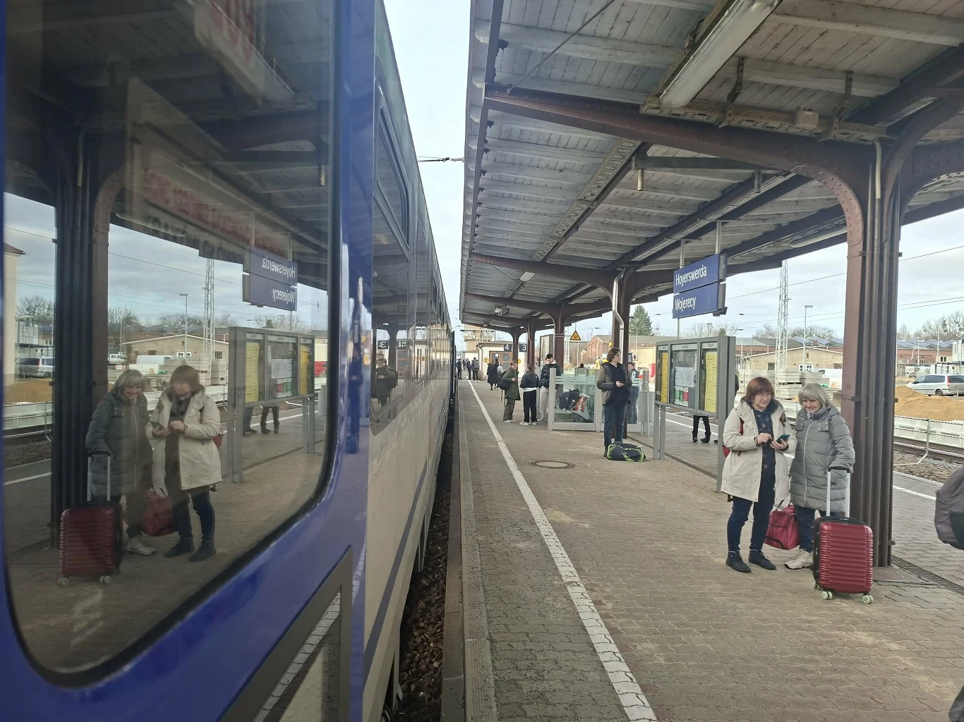 Polski pociąg zameldował się na stacji Hoyerswerda w Niemczech Peron dworca kolejowego z zatrzymanym niebieskim pociągiem, odbicie osób w oknie wagonu, pasażerowie z walizkami oczekują na peronie, zadaszenie i oznaczenie stacji w tle.