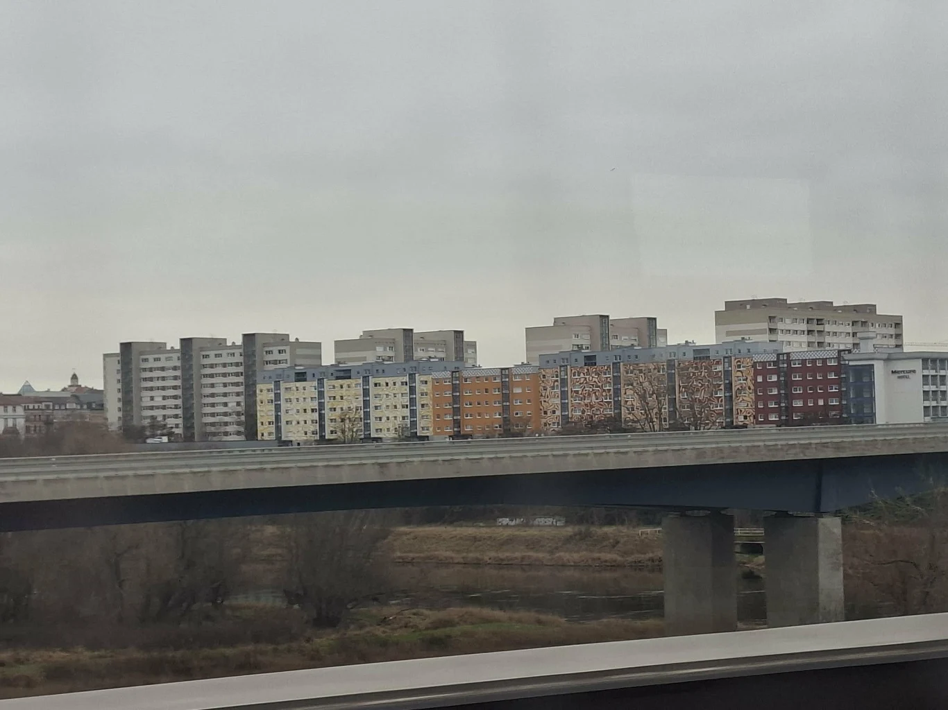 Mijana przez polski pociąg Riesa Szereg wysokich bloków mieszkalnych o różnych kolorach elewacji ustawionych w jednej linii, na pierwszym planie fragment nowoczesnego mostu przebiegającego nad suchym terenem lub wyschniętą roślinnością.