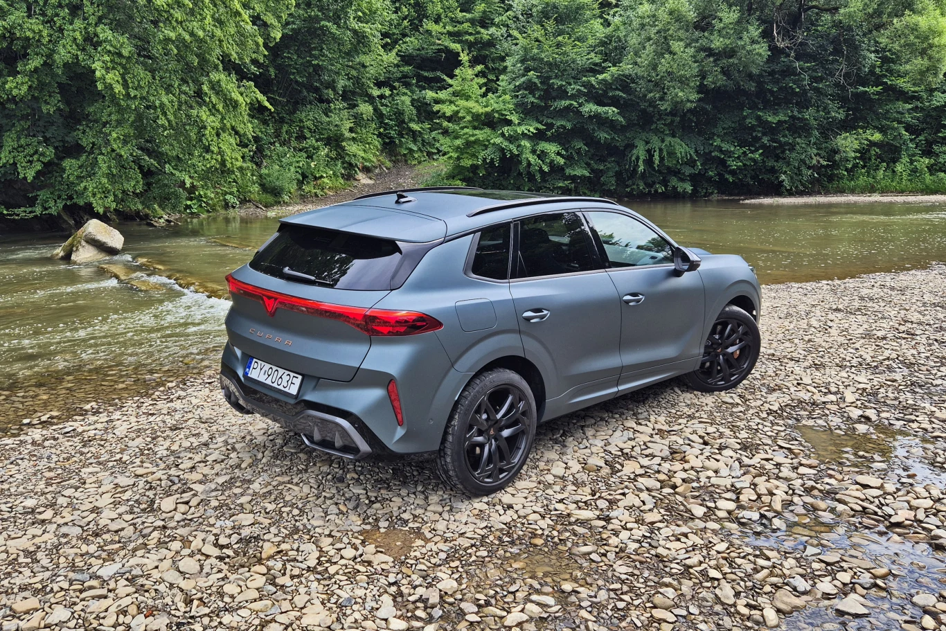 SUV z matowym szarym lakierem i czarnymi felgami zaparkowany na kamienistym brzegu przy rzece, w tle gęsty las i spokojna woda.