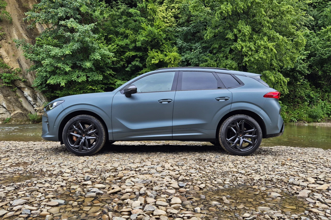 Nowoczesny, matowoniebieski SUV stoi na żwirowym nabrzeżu w otoczeniu bujnej zieleni i skał, z rzeką płynącą w tle.