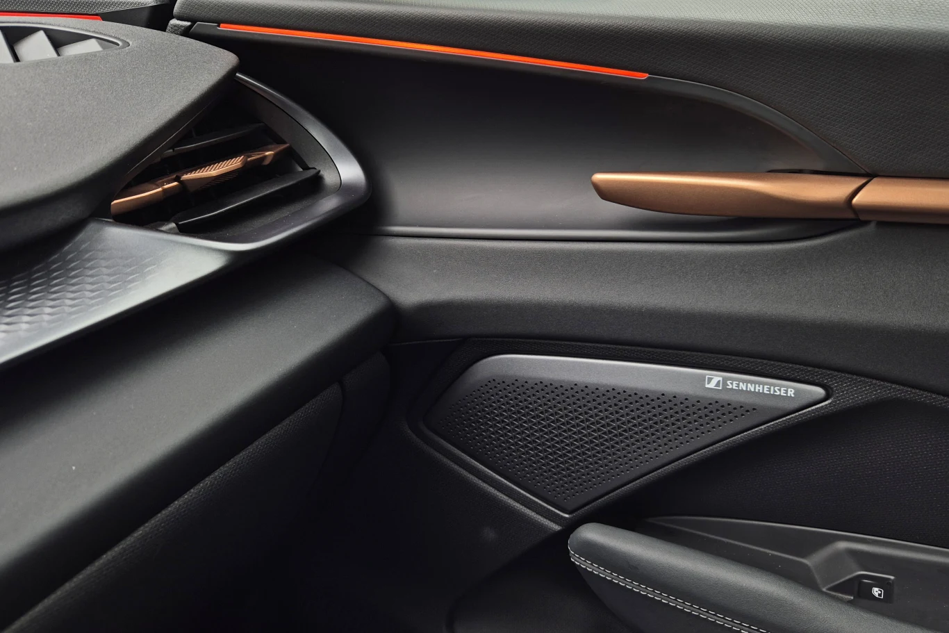 Fragment wnętrza samochodu z widocznym głośnikiem marki Sennheiser, stylowymi wykończeniami drzwi oraz akcentami w kolorze miedzi i pomarańczy.
