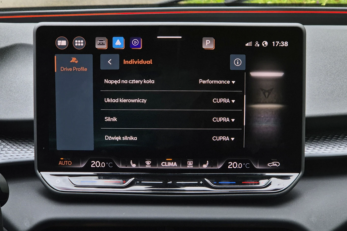 Centralny wyświetlacz samochodowy z menu ustawień trybu jazdy oraz klimatyzacji, widoczne ikony aplikacji na górnej krawędzi ekranu oraz szczegóły dotyczące parametrów pojazdu, takich jak napęd na cztery koła, układ kierowniczy, silnik i dźwięk silnika.
