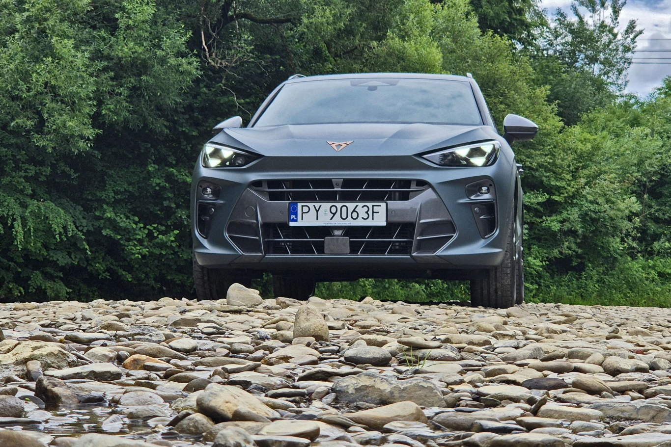 SUV marki Cupra zaparkowany na kamienistej drodze w otoczeniu zielonych drzew i krzewów, widoczny z przodu na wprost, z czytelną tablicą rejestracyjną.