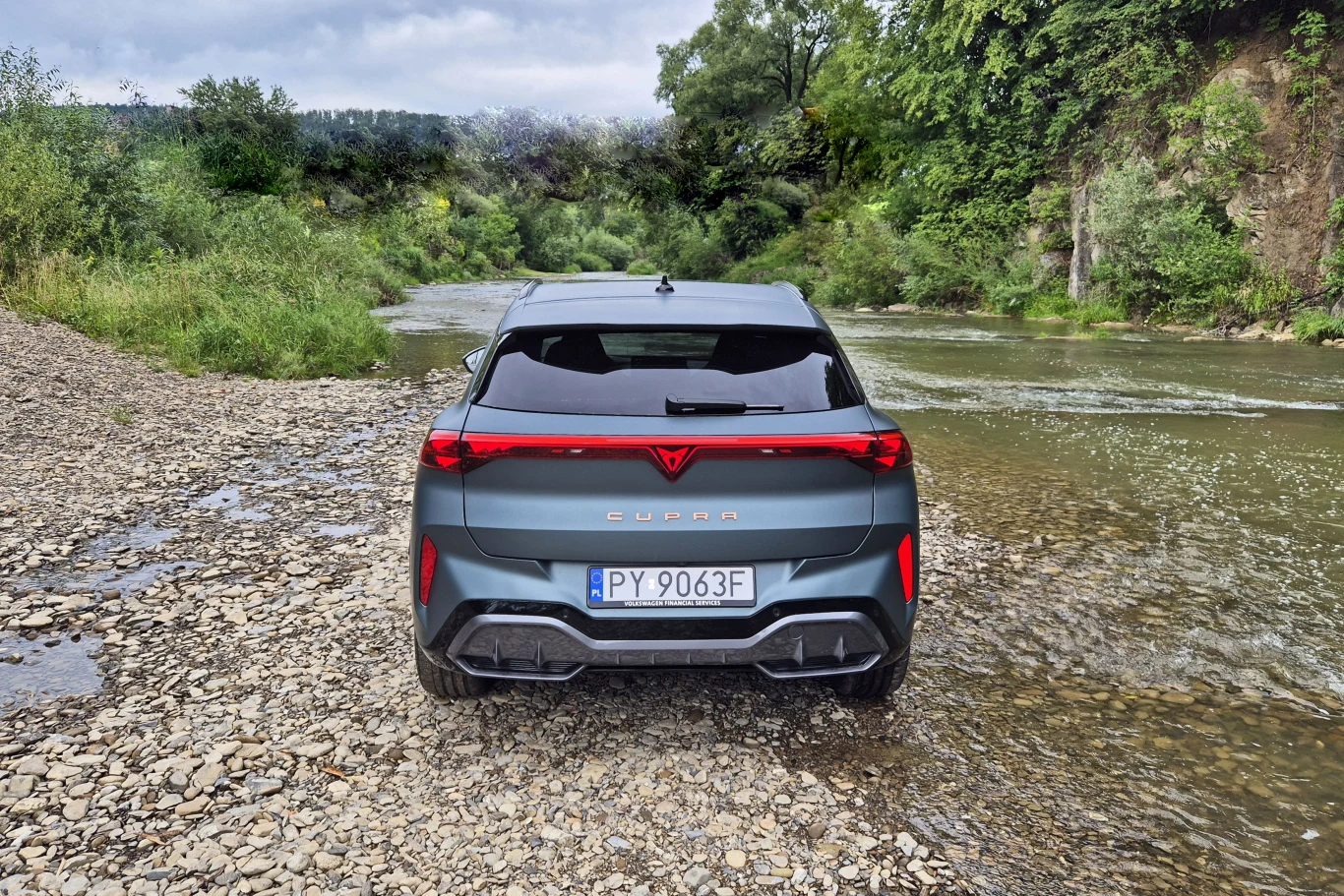 Samochód Cupra z polską tablicą rejestracyjną zaparkowany na kamienistym brzegu rzeki, otoczony zielenią i drzewami, w tle widać wodę oraz zalesione wzgórza.