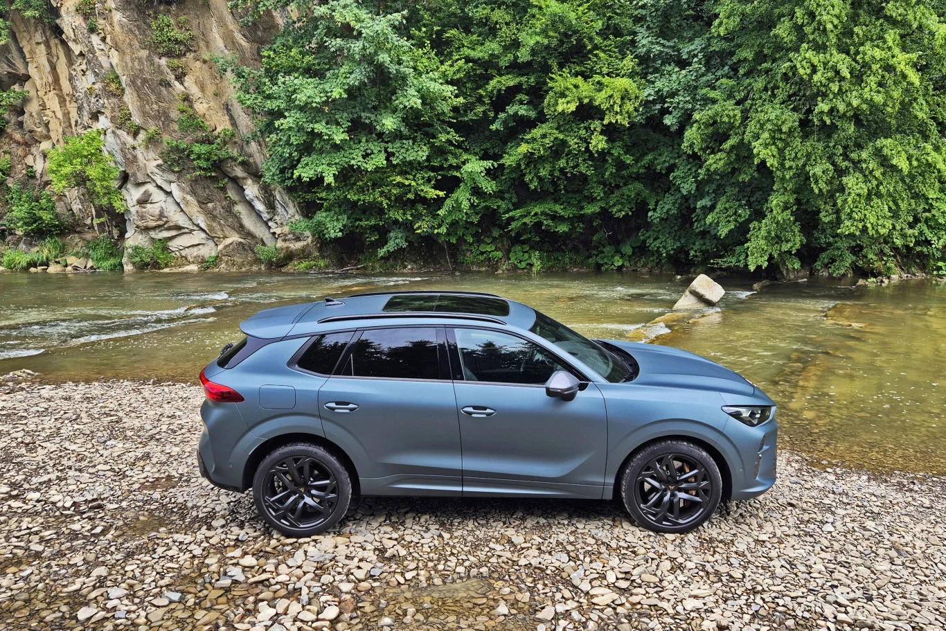Nowoczesny SUV w kolorze niebieskim zaparkowany na kamienistym brzegu rzeki, w tle gęste zielone drzewa i skaliste urwisko.