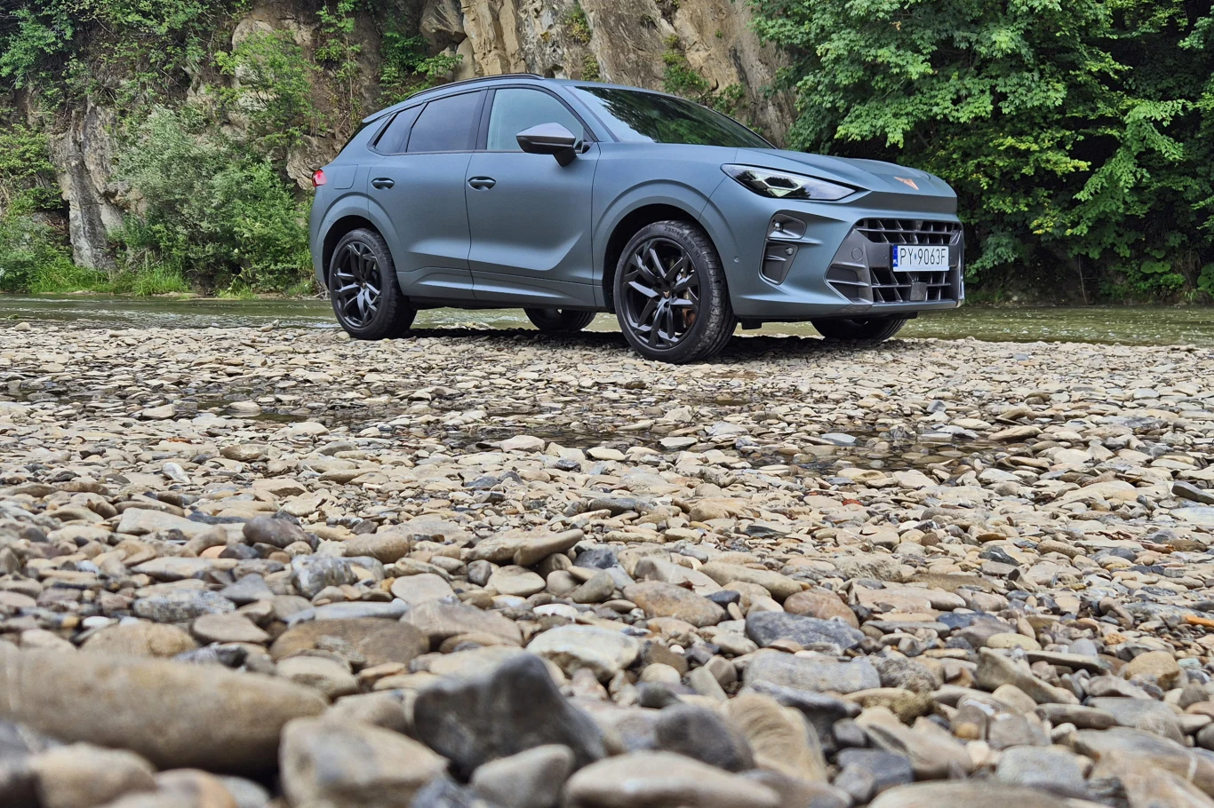 Nowoczesny, szary SUV stoi na kamienistym podwórzu, otoczony zielenią oraz stromymi skałami w tle, podkreślając terenowy charakter pojazdu.