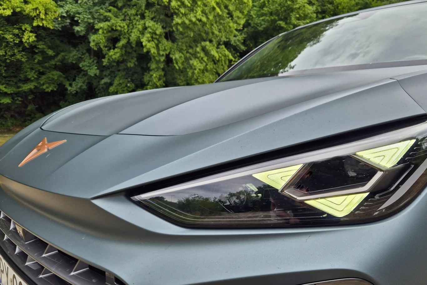 Nowoczesny samochód w kolorze matowego szarego, z charakterystycznym logo na masce oraz dynamicznie zaprojektowanym reflektorem, stojący na tle zielonych drzew.