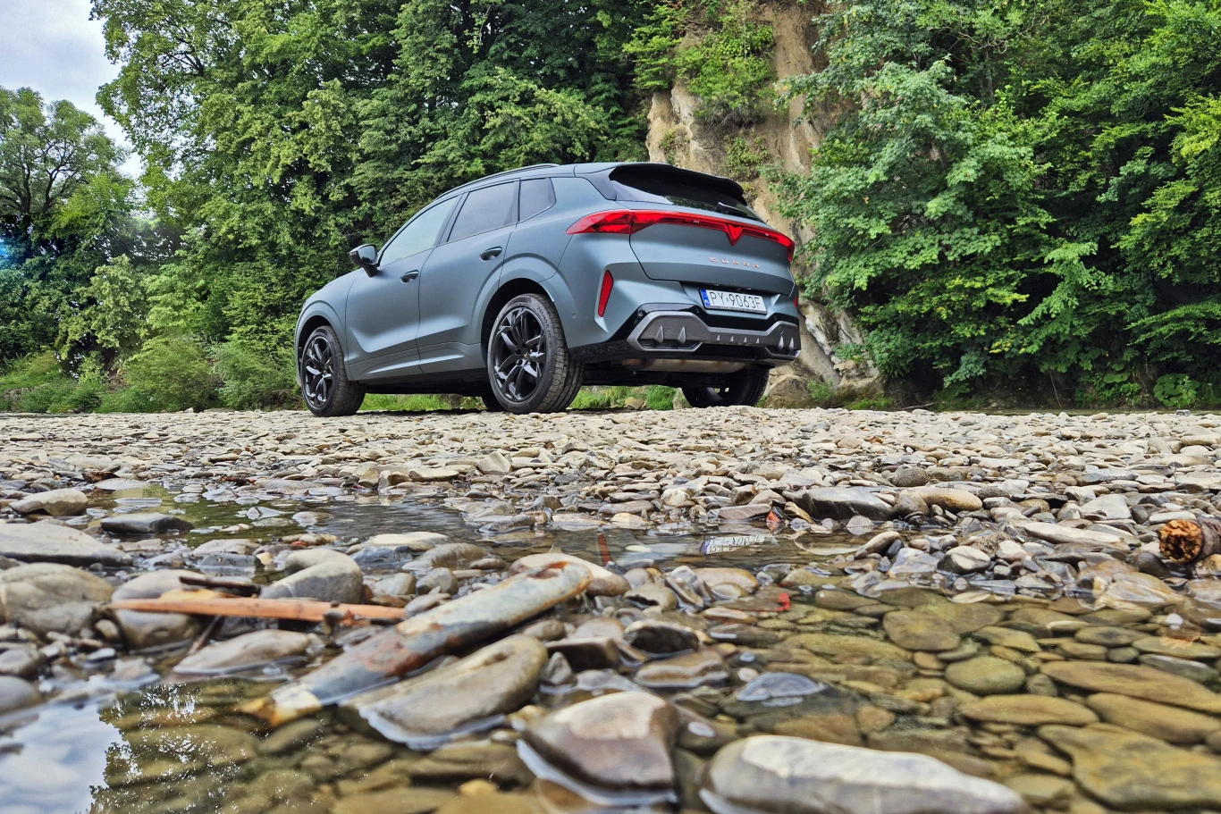 Nowoczesny samochód terenowy zaparkowany na żwirowym brzegu, w otoczeniu zielonych drzew i poszarpanych skał. Na pierwszym planie widać płytką wodę oraz kamienie, tworzące naturalny krajobraz.