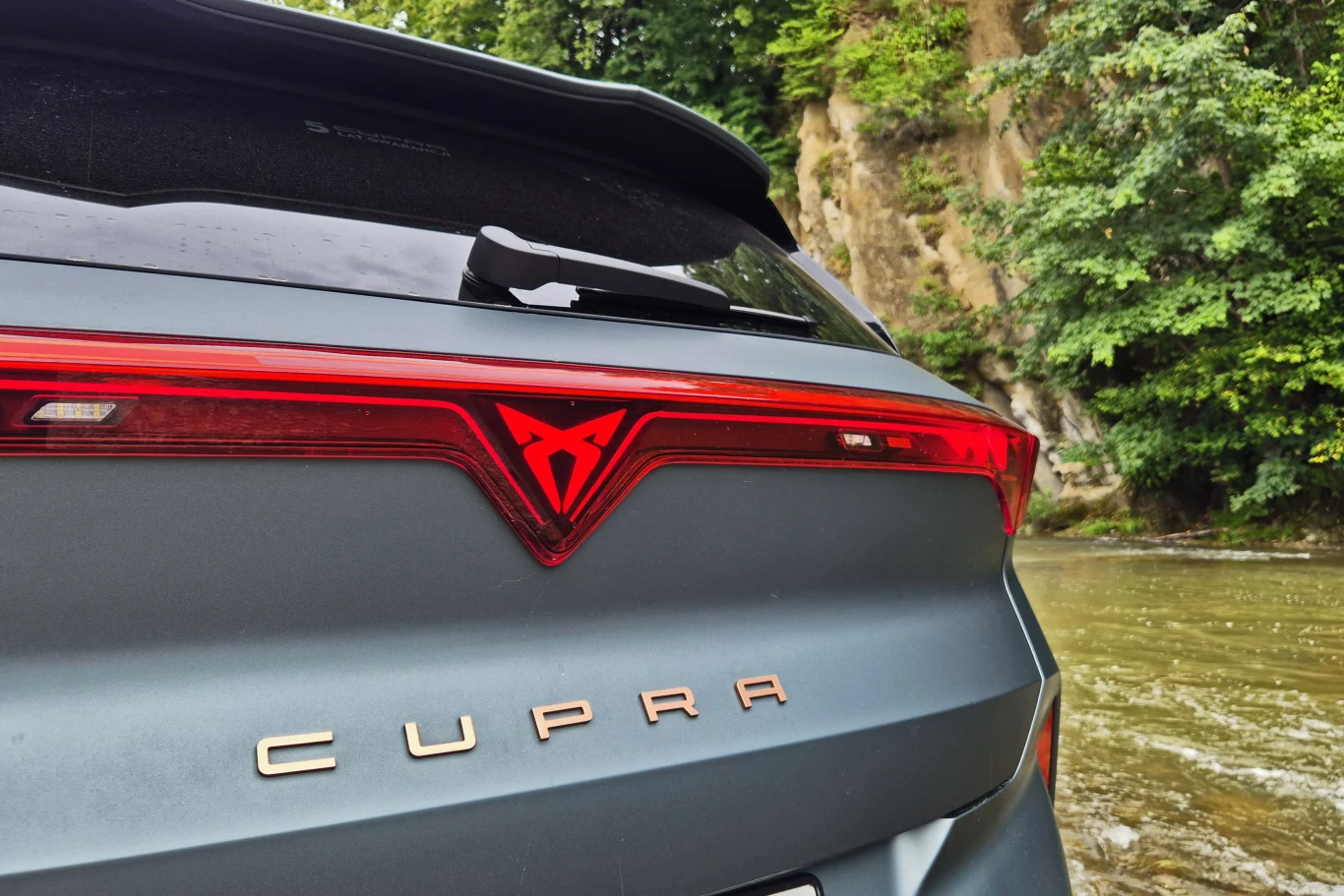 Tył samochodu marki Cupra z charakterystycznym logo na czerwonym pasku świateł, otoczony naturalnym krajobrazem z drzewami i rzeką w tle.