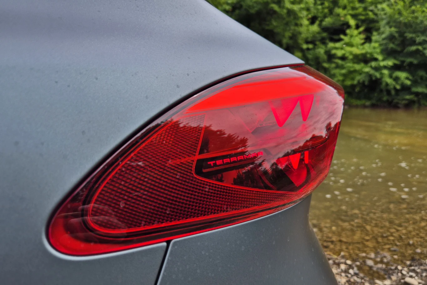 Czerwone światło tylnej lampy samochodowej na tle rozmytej zieleni i żwirowej powierzchni przy wodzie, na lampie widoczny napis Terra.