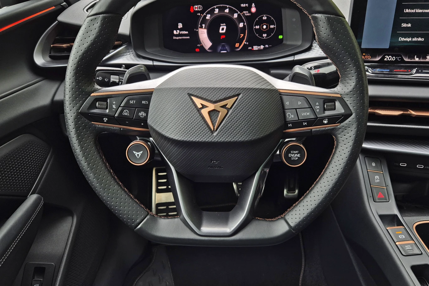 Sportowa kierownica z logo marki Cupra, liczne przyciski umożliwiające sterowanie systemami pojazdu, cyfrowy wyświetlacz z informacjami dla kierowcy, eleganckie wykończenia wnętrza samochodu oraz widoczne elementy deski rozdzielczej i konsoli centralnej.
