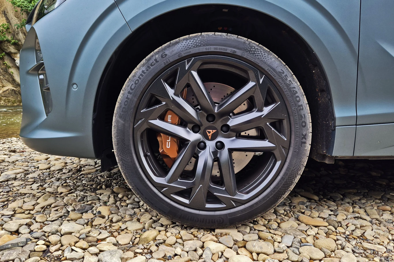 Duża, sportowa felga z charakterystycznym wzorem i widocznym zaciskiem hamulcowym, fragment karoserii samochodu SUV w matowym lakierze, pojazd stoi na kamienistym podłożu.