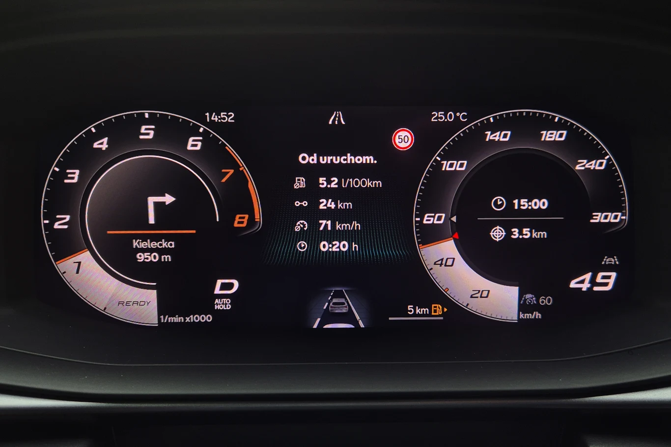 Cyfrowy kokpit samochodu z wyświetlaczem prędkościomierza, obrotomierza, informacjami o trasie, wskazówkami nawigacyjnymi oraz danymi o spalaniu paliwa. Centralnie wyświetlana mapa z instrukcjami jazdy oraz szczegółami dotyczącymi bieżącej podróży.