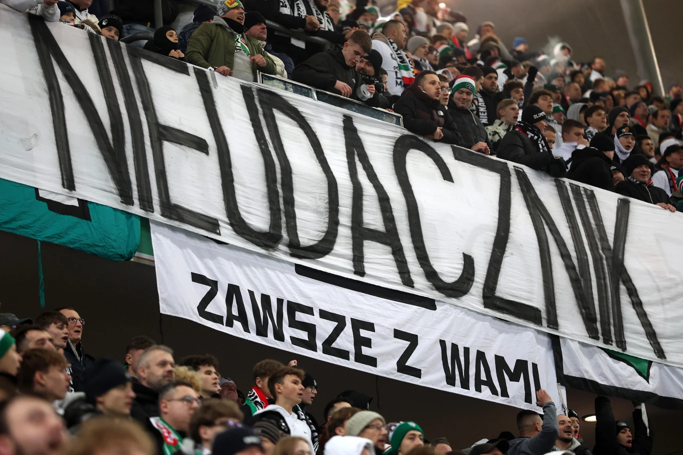 Tłum kibiców na stadionie, trzymających duży transparent z napisem 'NIEUDACZNIK' oraz mniejszy transparent 'ZAWSZE Z WAMI', wyrażających silne emocje wobec odbiorcy transparentu.
