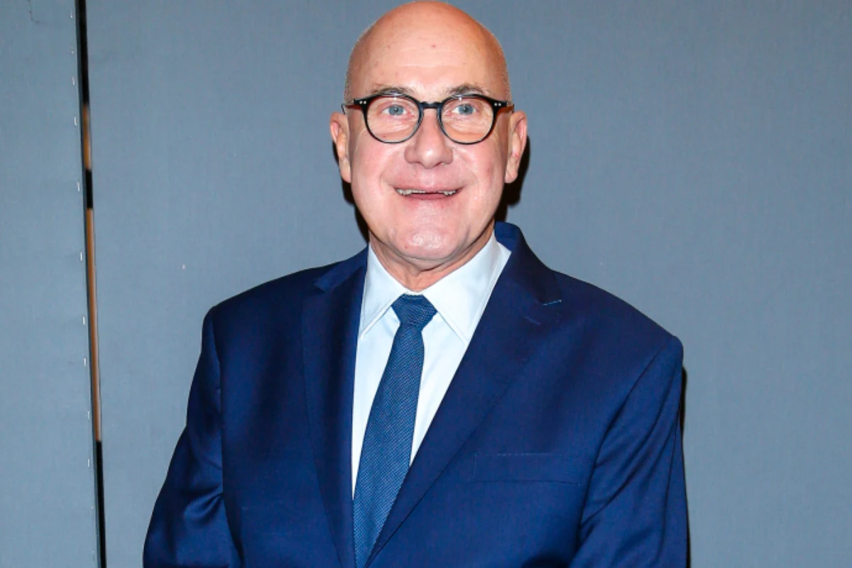 Piotr Machalica Mężczyzna w średnim wieku w granatowym garniturze, jasnoniebieskiej koszuli i granatowym krawacie stoi na tle szarej ściany, uśmiechnięty, w okularach.