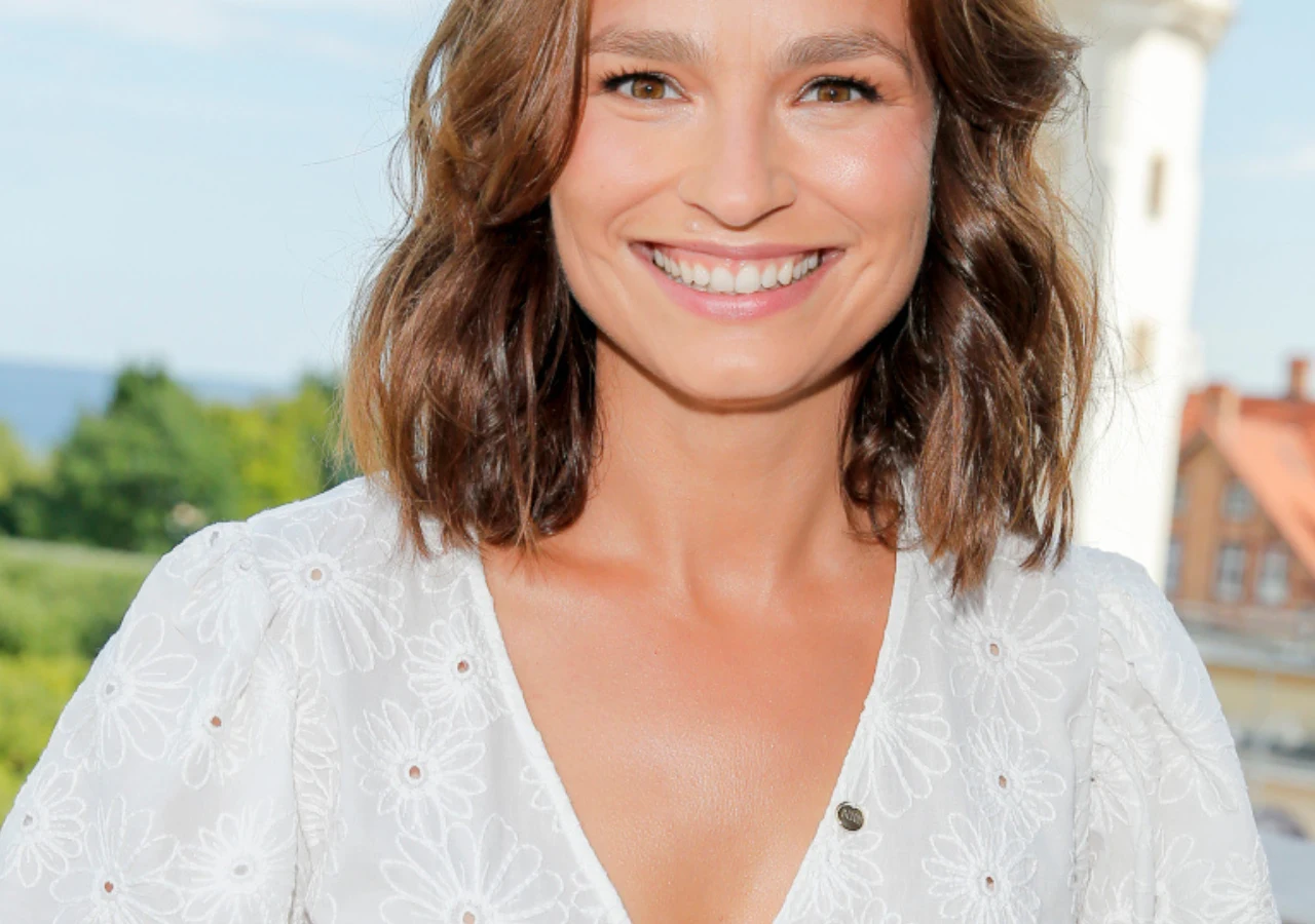 Anna Starmach Uśmiechnięta kobieta o brązowych włosach w białej sukience z koronkowym wzorem, stoi na tle rozmytego krajobrazu z zielenią i fragmentami architektury.