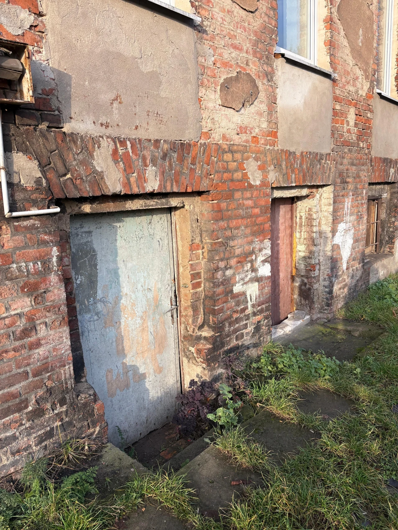 Zniszczona ceglana ściana starego budynku z widocznymi śladami łuszczącego się tynku. Dwa wejścia do piwnicy, jedno z metalowymi, zardzewiałymi drzwiami, drugie z drewnianymi, wyblakłymi drzwiami. Otoczenie porośnięte trawą i chwastami.