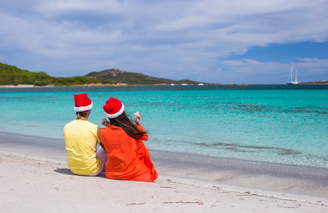Para w czapkach świątecznych siedzi na białym piasku patrząc na turkusowe morze, w tle zielone wzgórza i żaglówki na wodzie pod błękitnym niebem.