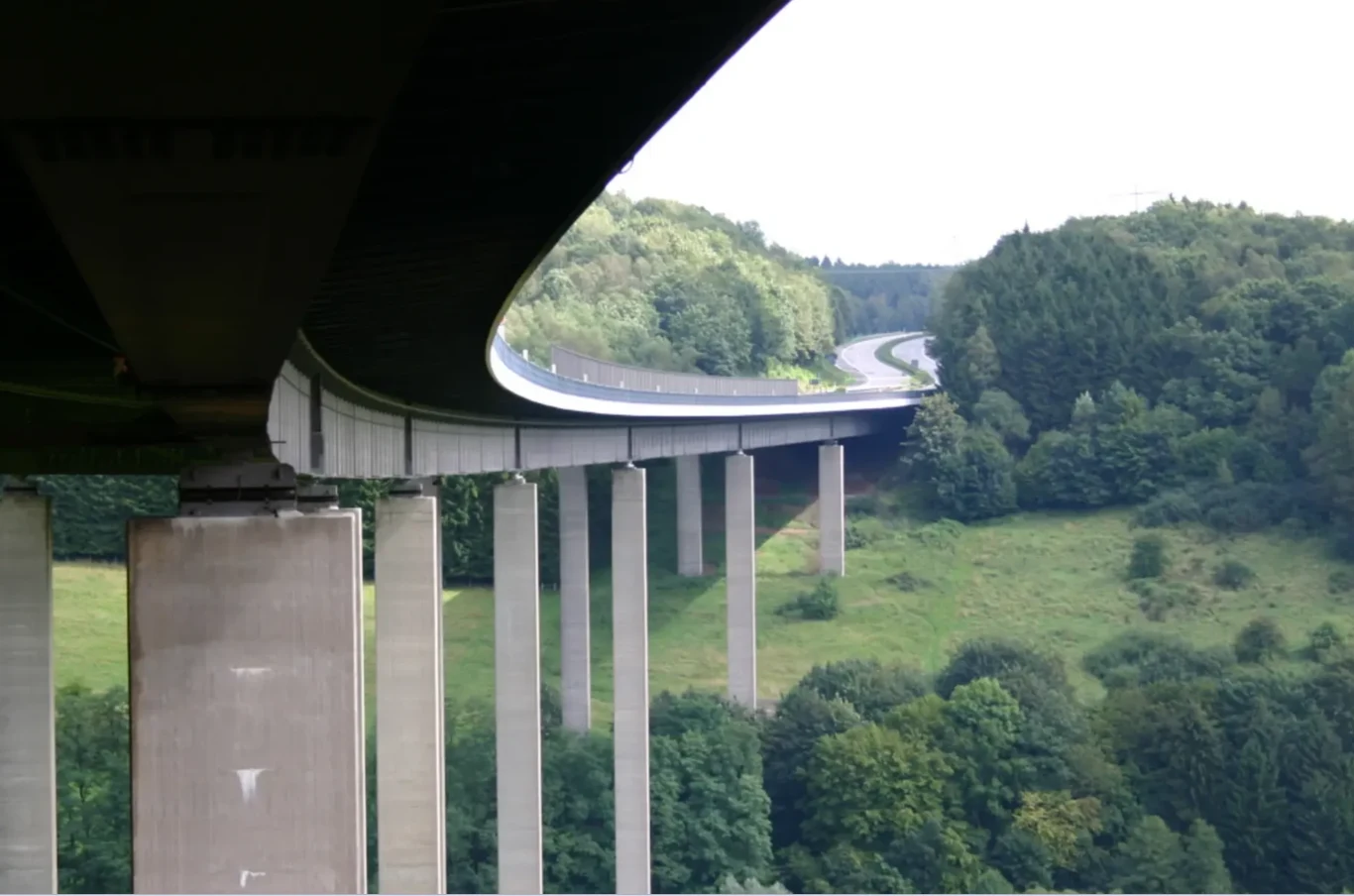 Most na wysokich filarach przerzucony nad zieloną doliną, w tle widoczne zalesione wzgórza oraz kręta droga prowadząca przez las.