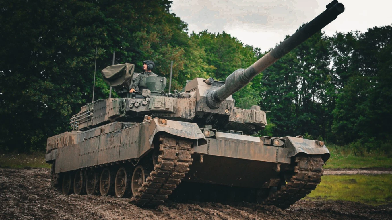 Nowoczesny czołg poruszający się po błotnistym terenie, otoczony gęstym lasem, z widocznym żołnierzem w otwartym włazie wieży.