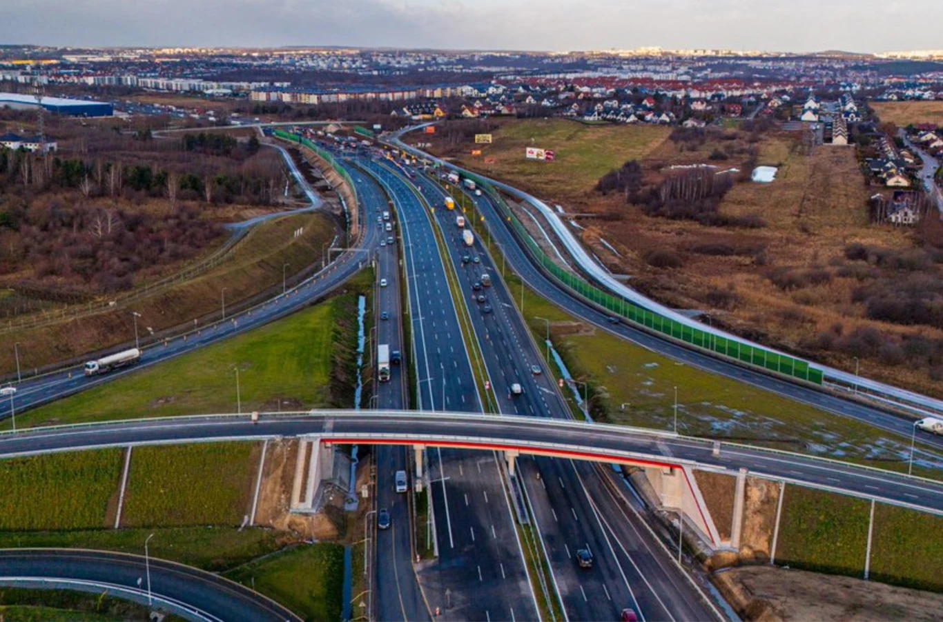 Obwodnica Gdańska z nowym układem jezdni. GDDKiA wprowadza zmiany. Nowoczesna autostrada otoczona ekranami dźwiękochłonnymi, z widocznymi samochodami i ciężarówkami na kilku pasach ruchu, nad drogą przebiega most prowadzący nad trasą, w tle rozciągają się zabudowania mieszkalne oraz tereny zielone.