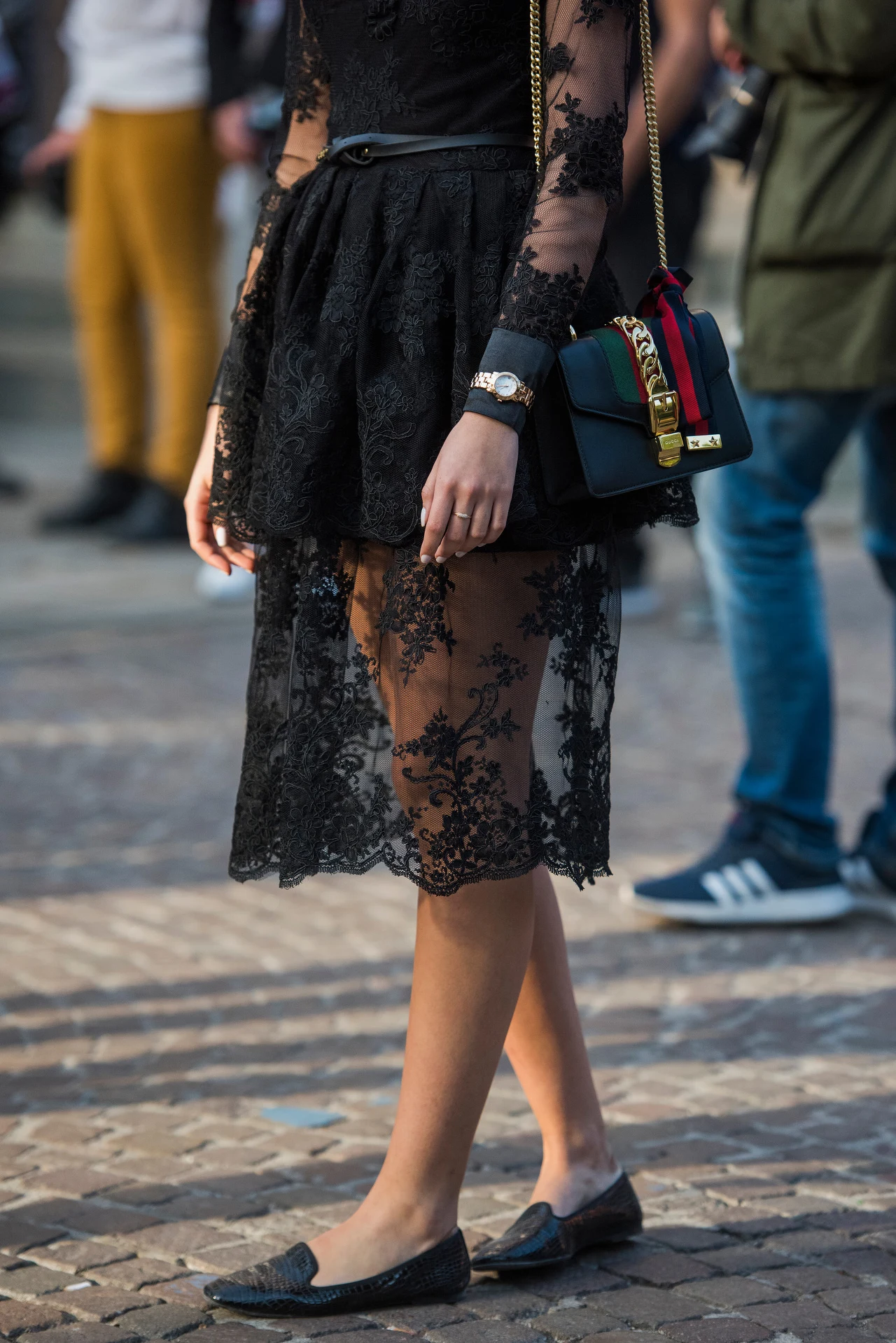 Kobieta ubrana w elegancką, czarną koronkową sukienkę z przezroczystym dołem i długimi rękawami, na ramieniu modna torebka na łańcuszku w kolorze czarnym z detalami, na nogach czarne baleriny, w tle rozmazane sylwetki przechodniów.