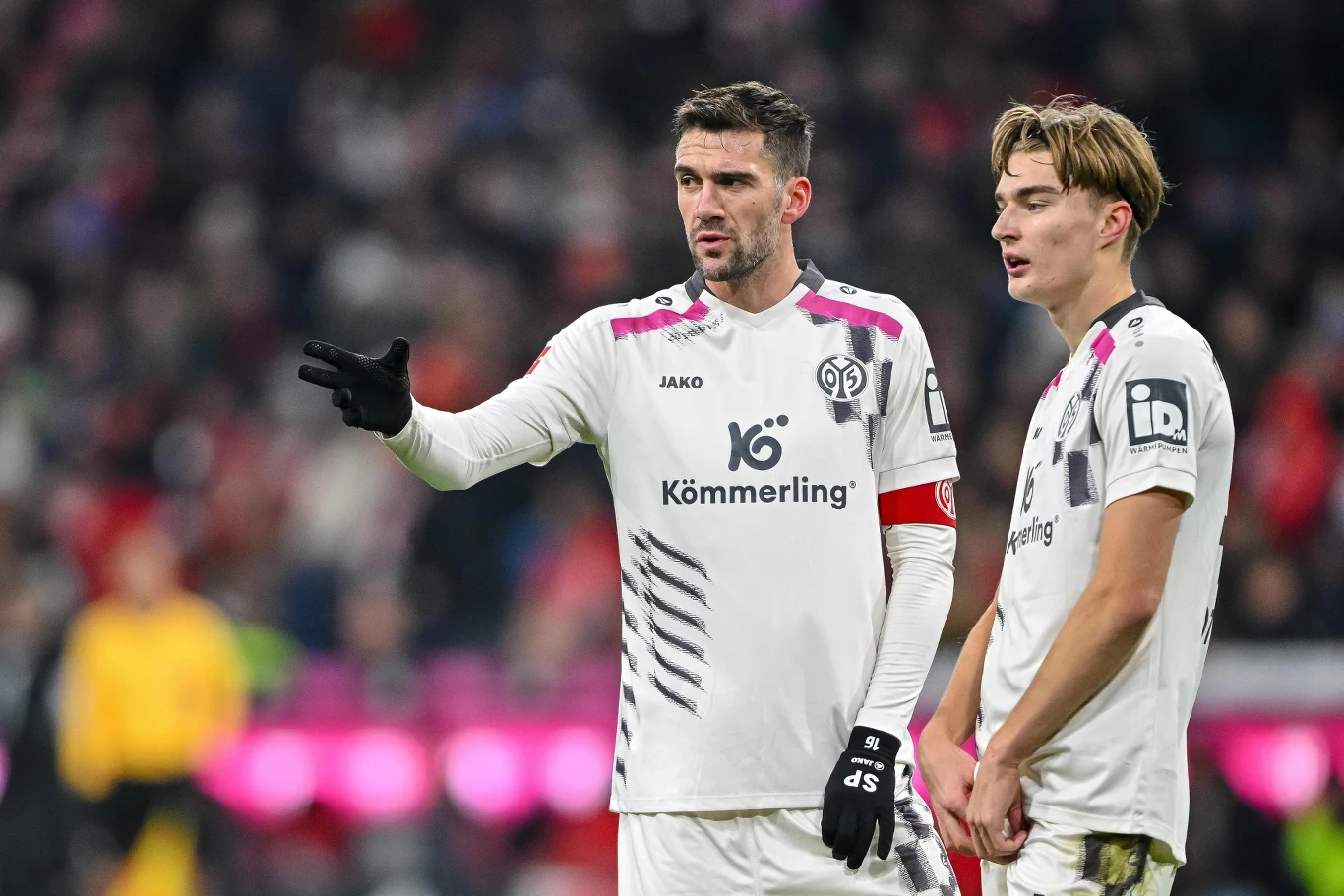 Kacper Potulski (z prawej) Dwóch piłkarzy drużyny FSV Mainz 05 w białych strojach sportowych rozmawia na boisku podczas meczu piłkarskiego, jeden z nich pokazuje ręką kierunek lub wydaje instrukcje, wokół widoczny rozmazany tłum kibiców na trybunach.