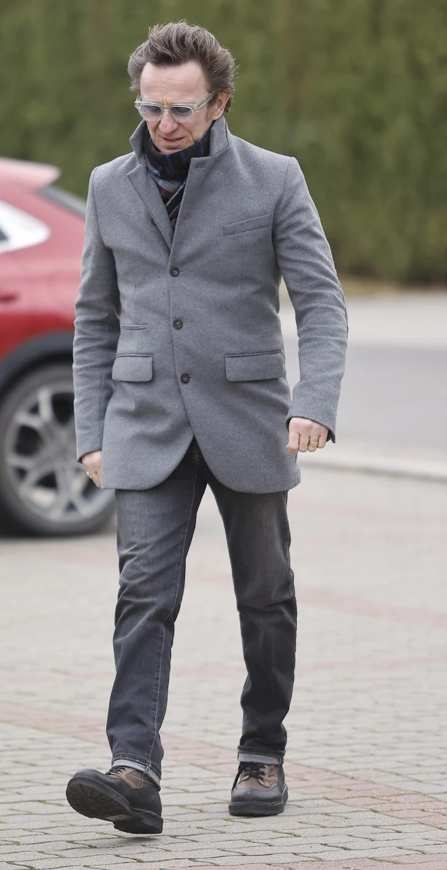 Paweł Wilczak na pogrzebie Agnieszki Maciąg Mężczyzna w szarym płaszczu i ciemnych spodniach spaceruje chodnikiem, w tle widoczny fragment czerwonego samochodu oraz zielony żywopłot.