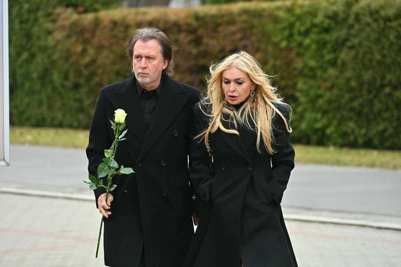 Tomasz Ziółkowski i Monika Olejnik na pogrzebie Agnieszki Maciąg Dwoje osób ubranych na czarno idzie obok siebie chodnikiem, jedna z nich trzyma białą różę. W tle widoczny zielony żywopłot i fragment ulicy. Oboje mają poważne wyrazy twarzy.