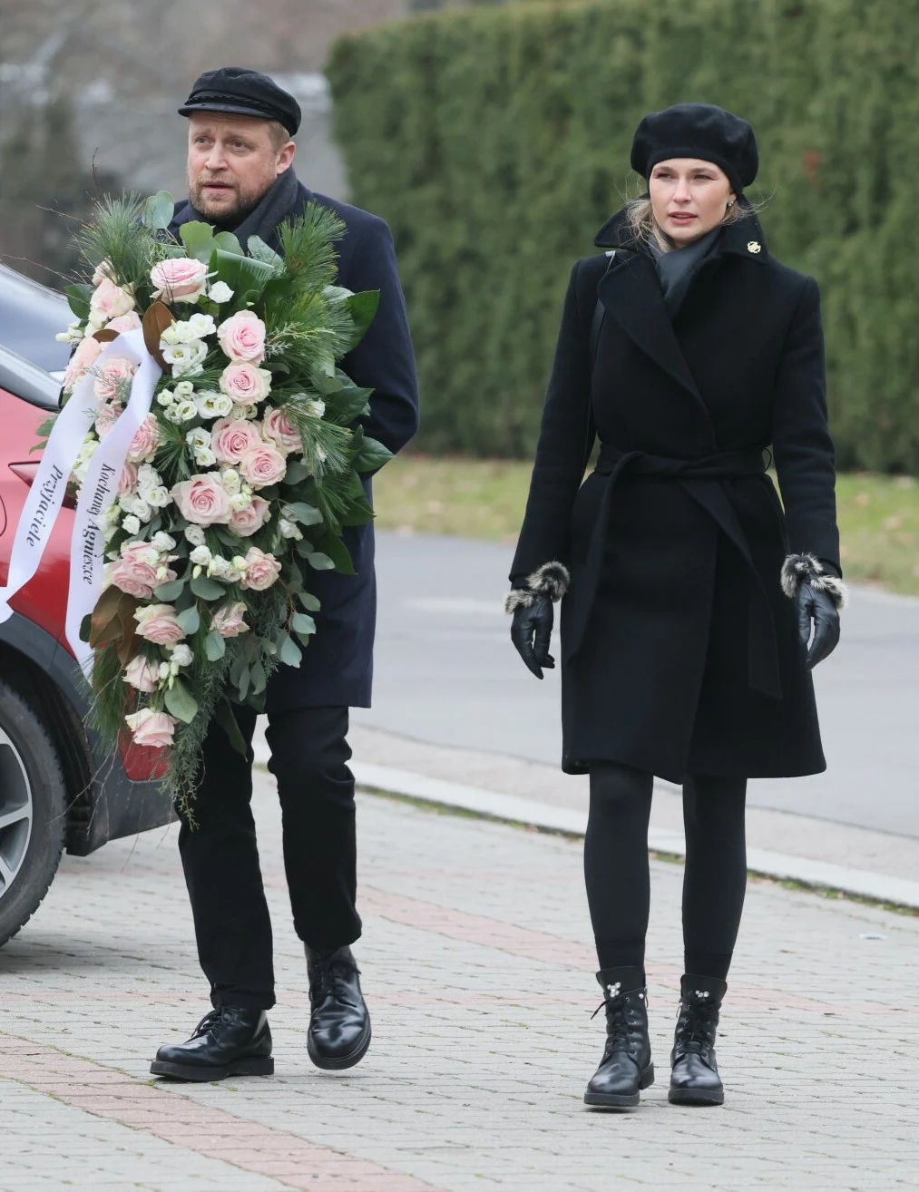Piotr Adamczyk, Karolina Szymczak na pogrzebie Agnieszki Maciąg Mężczyzna i kobieta ubrani na czarno idą chodnikiem, mężczyzna niesie dużą wiązankę z różowych i białych kwiatów oraz zielonych liści, w tle widoczne auto i zielony żywopłot.