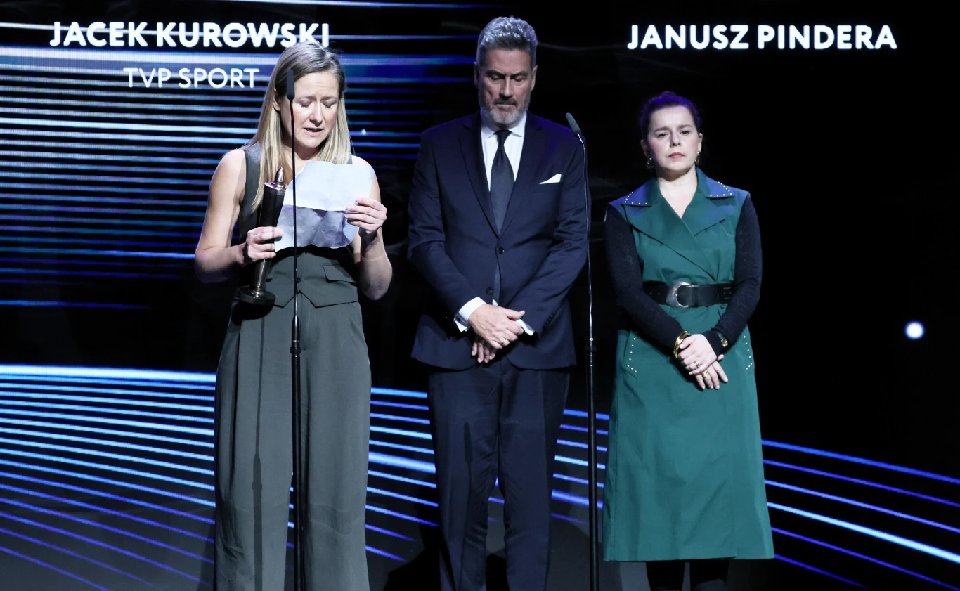 Trzy osoby stoją na scenie w świetle reflektorów, kobieta po lewej czyta coś z kartki, mężczyzna i druga kobieta patrzą w dół. W tle widoczne są nazwiska Jacek Kurowski TVP Sport oraz Janusz Pindera na czarnym tle z białymi napisami.
