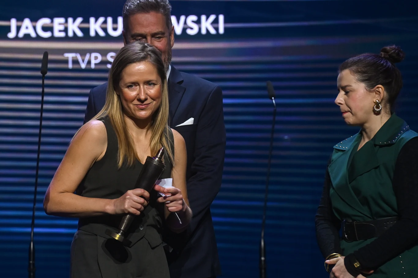 Kobieta w eleganckiej sukni trzymająca statuetkę nagrody podczas uroczystej gali, obok niej stoją dwie osoby na tle niebieskiej scenografii z wyświetlonym napisem.