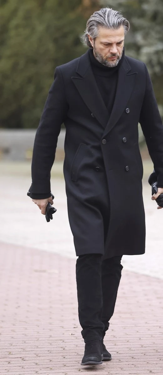 Olivier Janiak na pogrzebie Agnieszki Maciąg Mężczyzna o siwych włosach i brodzie idący chodnikiem w eleganckim, ciemnym płaszczu i czarnych ubraniach, trzyma w rękach czarne rękawiczki. Ujęcie plenerowe z rozmytym tłem zieleni.