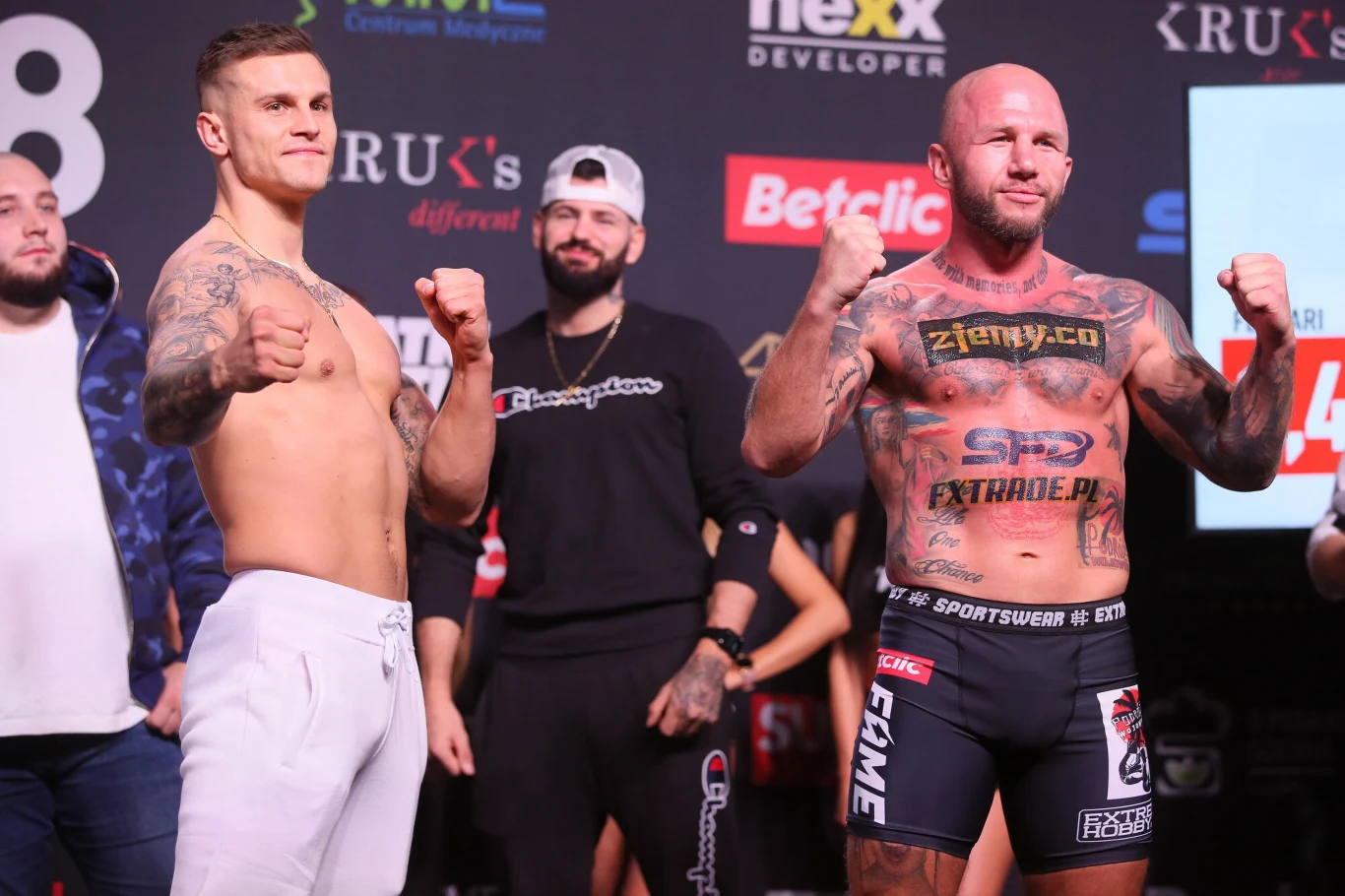 Dwóch umięśnionych mężczyzn w sportowych spodniach stoi z zaciśniętymi pięściami naprzeciwko siebie na tle sponsorów i banerów, otoczeni innymi osobami, atmosfera sportowej rywalizacji przed walką.