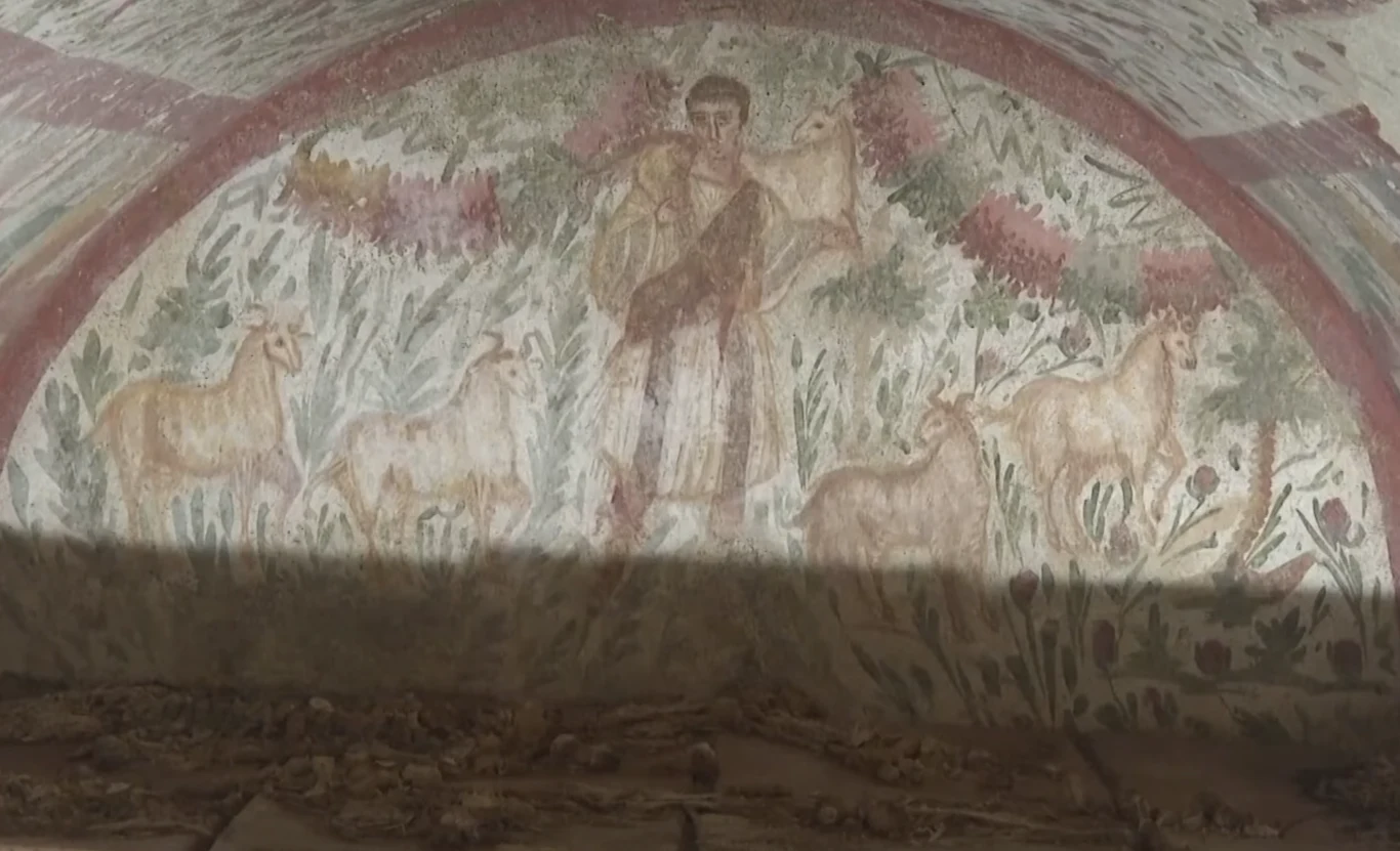 Ścienna malowidło przedstawiające postać otoczoną przez sześć owiec na tle zieleni i elementów roślinnych, całość utrzymana w kolorystyce pastelowej, część dolna obrazu jest częściowo zasłonięta cieniem.