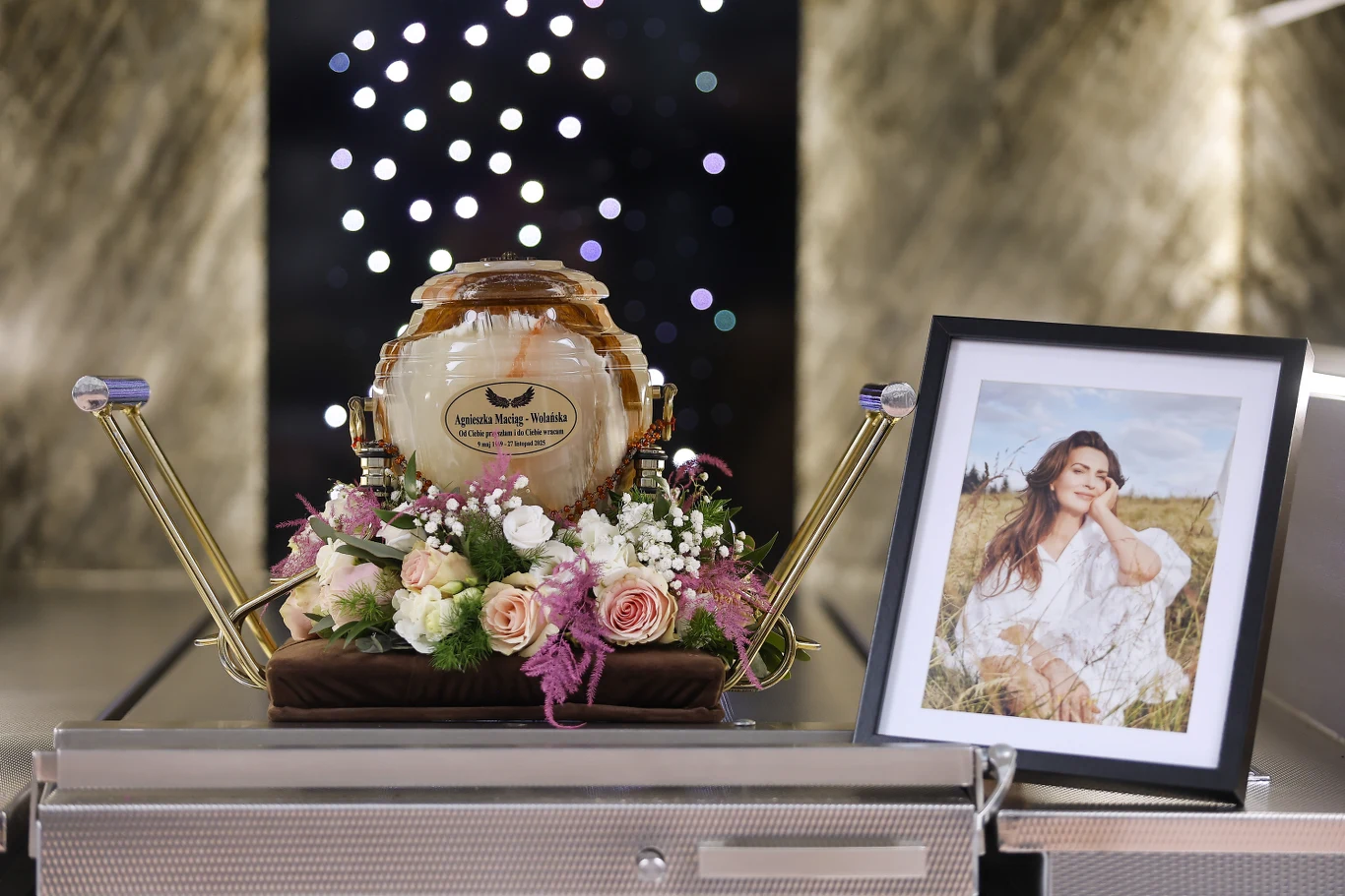 Urna ozdobiona bukietem kwiatów, obok ustawione oprawione zdjęcie uśmiechniętej kobiety na tle natury, całość ustawiona na kremacyjnym wózku w nowoczesnym pomieszczeniu.