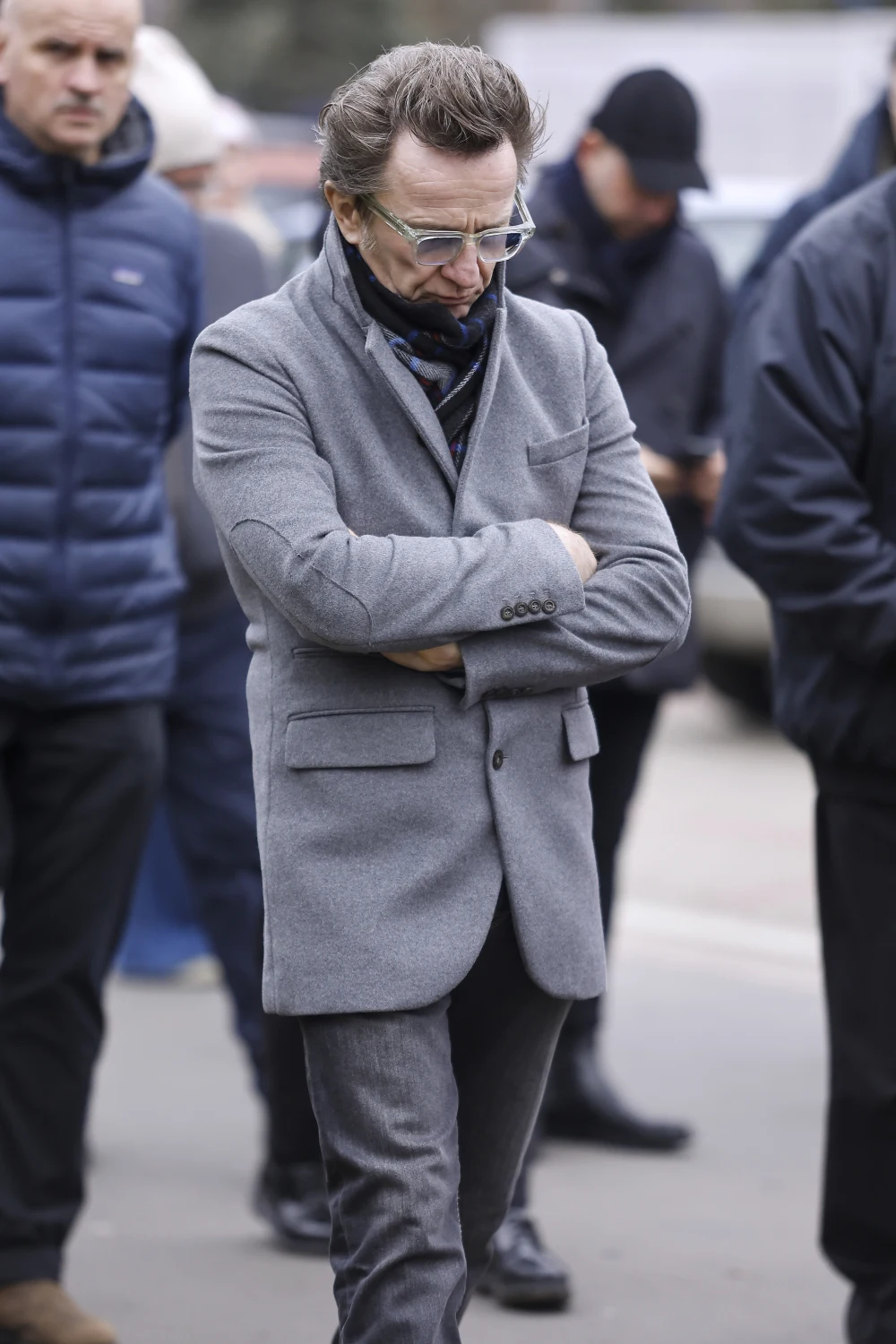 Mężczyzna w okularach, ubrany w szary garnitur i czarny szalik, idzie ze skrzyżowanymi ramionami, sprawiając wrażenie zamyślonego lub zamkniętego w sobie. W tle widoczni są inni ludzie w ciemnych kurtkach, sceneria miejska.