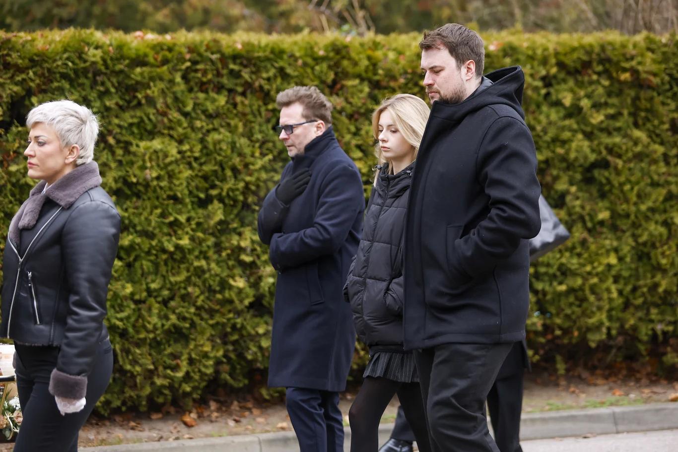 Smętna grupa osób ubranych na ciemno idąca alejką, na tle zielonego żywopłotu, atmosfera wyraźnie poważna i refleksyjna, wyraz twarzy sugeruje żałobę lub udział w uroczystości pogrzebowej.