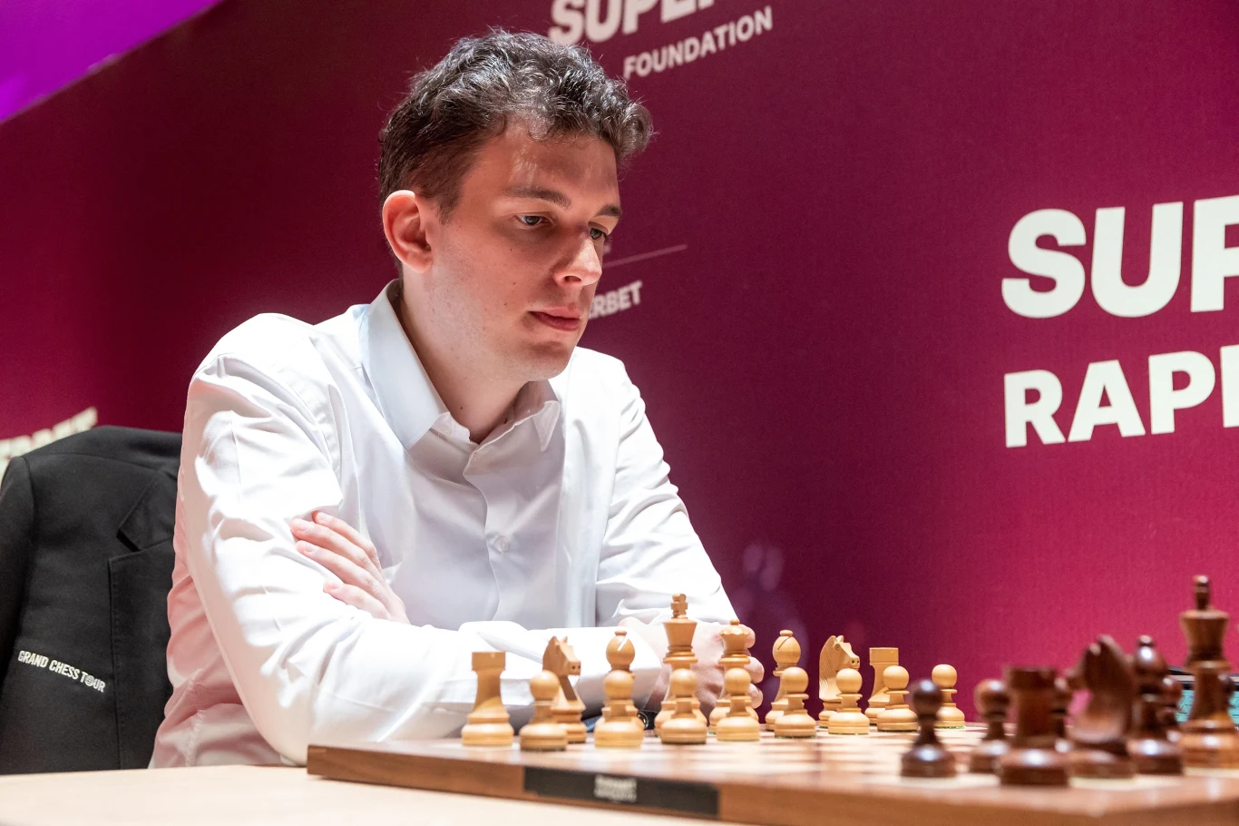 Mężczyzna w białej koszuli skupiony nad szachownicą podczas turnieju, z widocznymi figurami szachowymi i tłem z logo turnieju Superbet Rapid. Jasne oświetlenie i profesjonalna atmosfera podkreślają rangę wydarzenia.