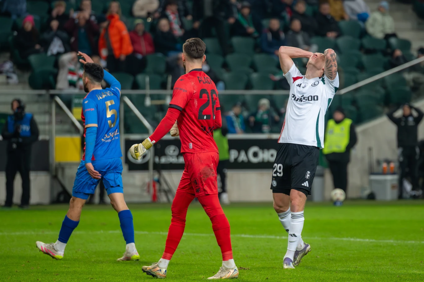 Mileta Rajović Piłkarze na boisku podczas meczu, jeden z zawodników w białej koszulce trzyma ręce na głowie, wyrażając rozczarowanie, bramkarz w czerwonym stroju oraz inny zawodnik drużyny przeciwnej odwróceni plecami. Kibice na trybunach w tle.