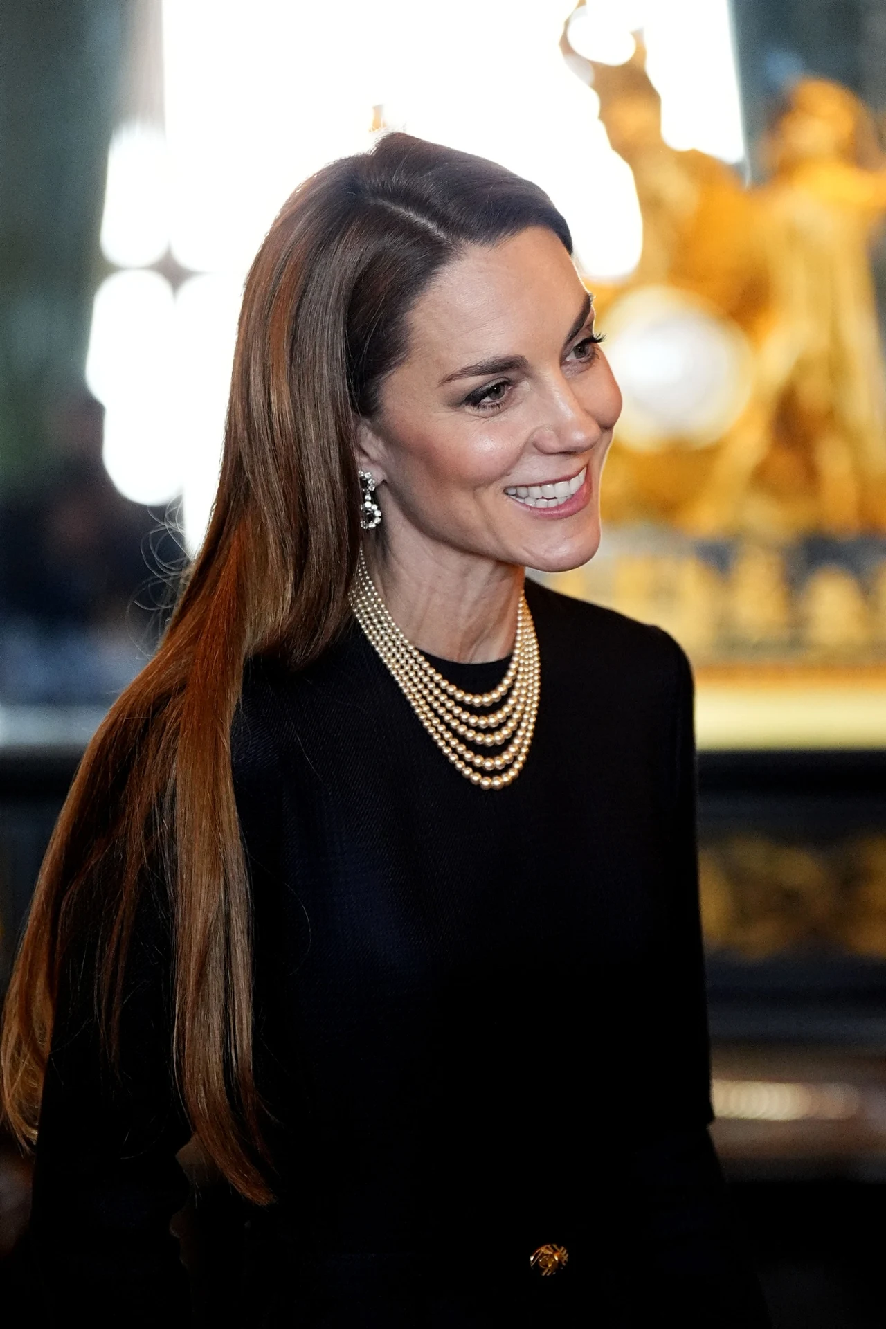 Elegancka kobieta o długich, prostych włosach ubrana w czarną suknię z perłową kolią i kolczykami, uśmiechnięta, pozująca w bogato zdobionym wnętrzu z dekoracyjną złotą rzeźbą w tle.