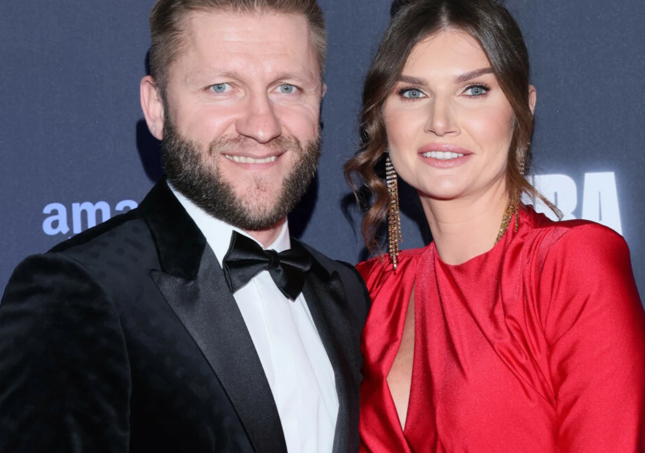 Jakub i Agata Błaszczykowscy Uśmiechnięty mężczyzna w czarnym smokingu i kobieta w czerwonej, eleganckiej sukni stoją razem na tle ścianki z logo wydarzenia, tworząc formalny, reprezentacyjny wizerunek pary.