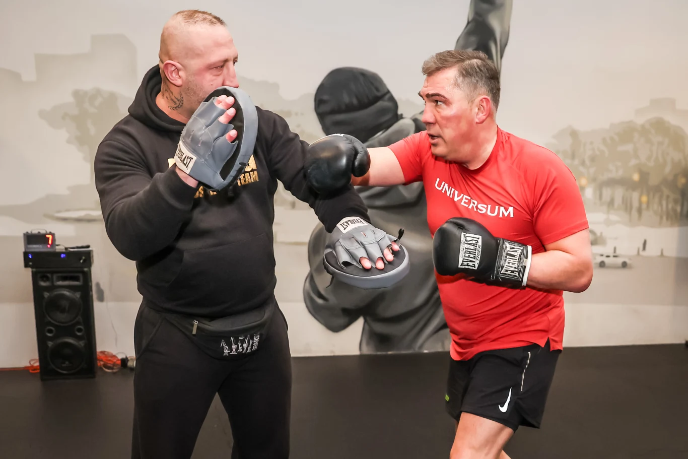 Dwóch mężczyzn podczas treningu bokserskiego, jeden z nich ubrany w czerwoną koszulkę wykonuje cios na tarcze trzymane przez drugiego mężczyznę w czarnym stroju i rękawicach. W tle widoczna ściana z muralem.