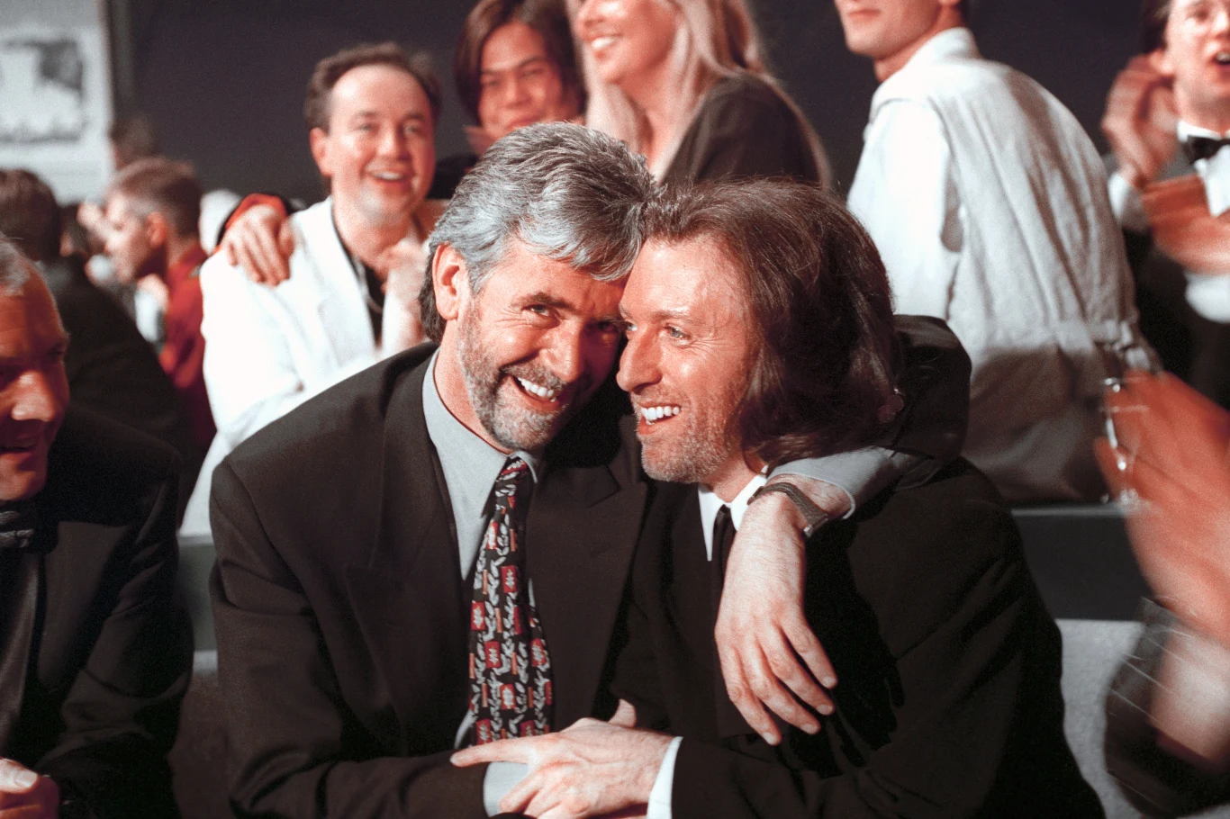 Charlie McGettigan (z lewej) i Paul Harrington po wygraniu Eurowizji 1994 w Dublinie (Irlandia) Dwóch uśmiechniętych mężczyzn ubranych elegancko obejmuje się i ściska w serdecznym geście, wokół nich śmiech i radość wśród tłumu ludzi ubranych formalnie.