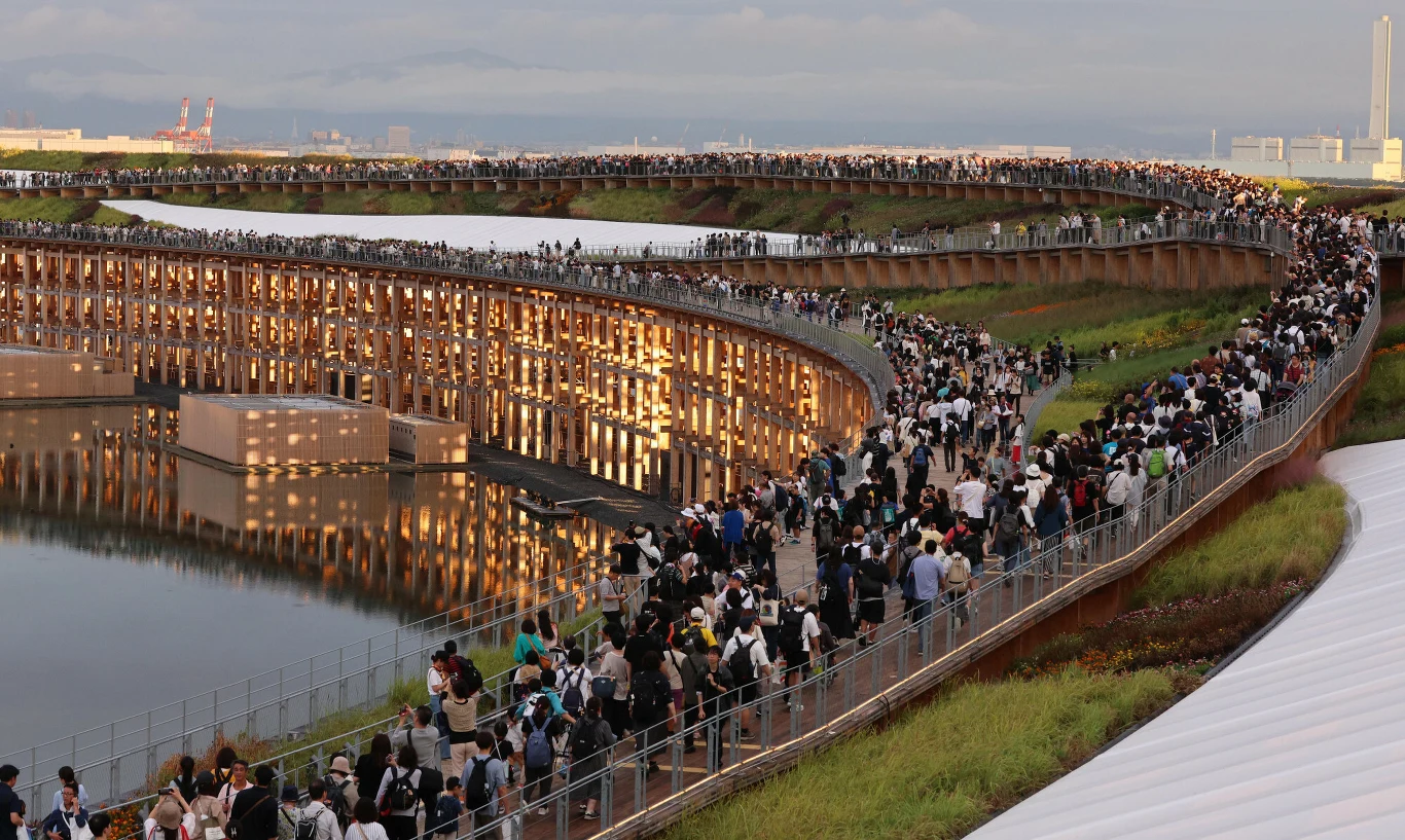 Tłum ludzi przemieszczający się po szerokiej krętej kładce nad wodą, pod którą widać podświetloną architekturę i zielone tereny po obu stronach mostu.