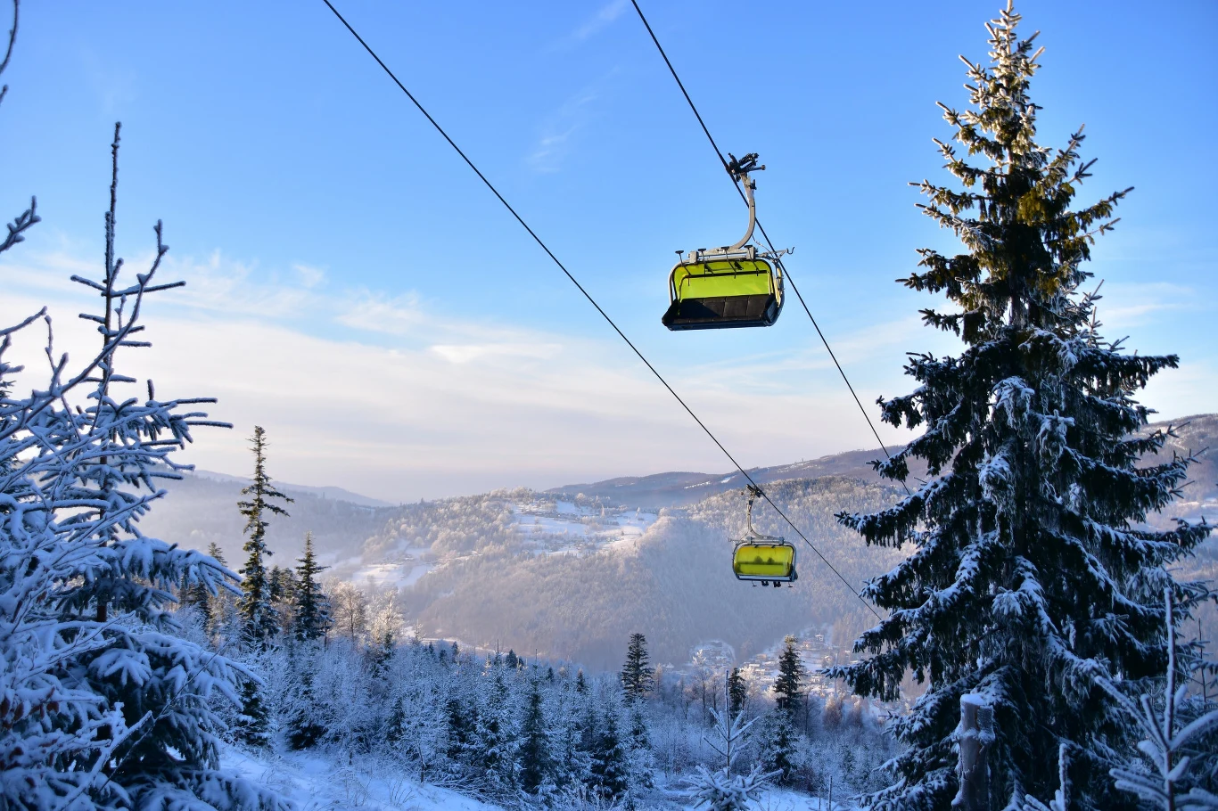Kolejka linowa z żółtymi wagonikami poruszająca się nad pokrytym śniegiem lasem iglastym, w tle widoczne górzyste krajobrazy oraz niebo o poranku lub zachodzie.