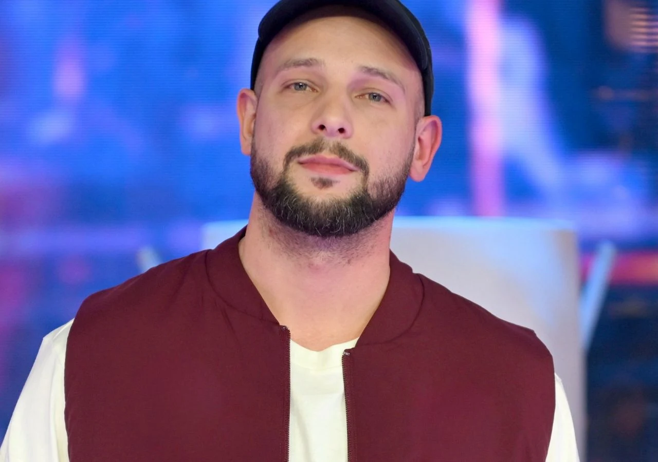 Maciej Pela Maciej Pela ubrany w bordową kurtkę, białą koszulkę i czarną czapkę z daszkiem na niebiesko-fioletowym tle.